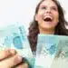 Mulher sorrindo e segurando várias notas de R$ 100, representando a alegria de receber o PIX ou pagamento na conta nesta quarta-feira (15).