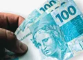 Mão segurando notas de R$ 100, simbolizando o último pagamento do Auxílio Gás em dinheiro em outubro de 2025.