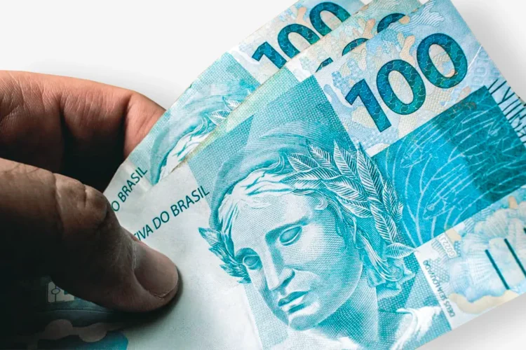Mão segurando notas de R$ 100, simbolizando o último pagamento do Auxílio Gás em dinheiro em outubro de 2025.