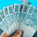 Datas de pagamento de outubro para Pé-de-Meia e Bolsa Família; veja o calendário 11 Mão segurando várias notas de 100 reais brasileiras em leque com fundo azul e notas caindo ao redor