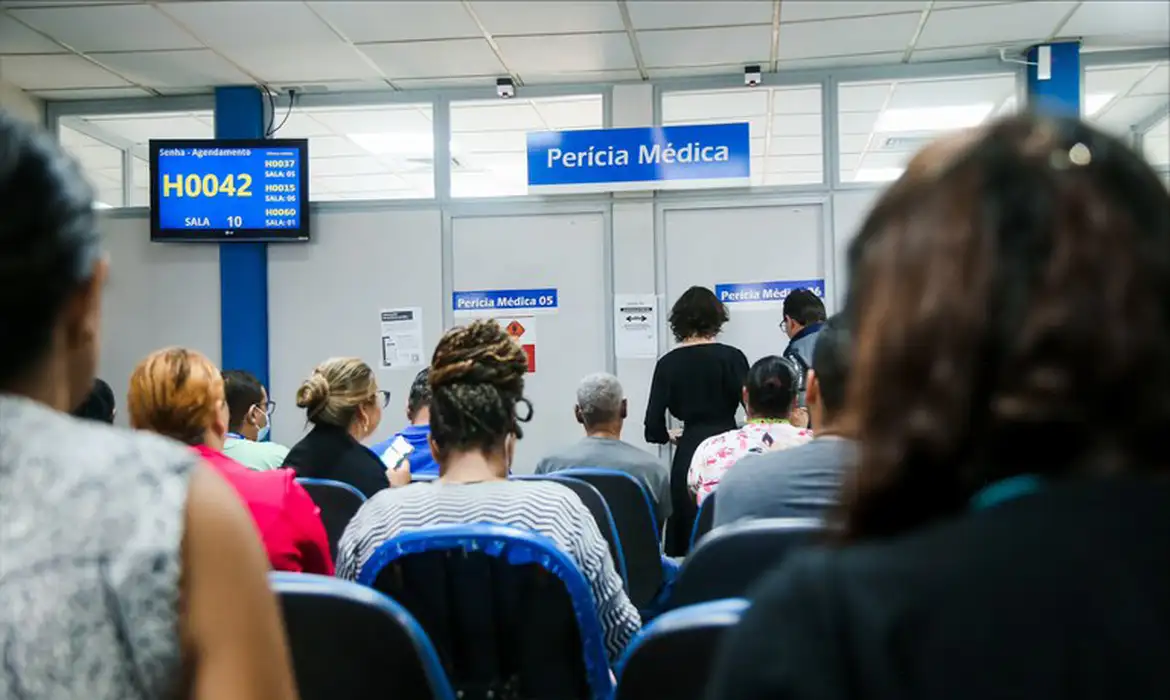 Imagem de uma sala de espera do INSS com a sinalização "Perícia Médica" e um painel de senhas, ilustrando o local onde serão realizados os atendimentos agendados nos dias 04 e 05 de outubro.