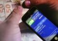 Mãos segurando um smartphone com o aplicativo Auxílio Emergencial da Caixa e cédulas de 50 reais, ilustrando o tema de devolução de valores indevidos.