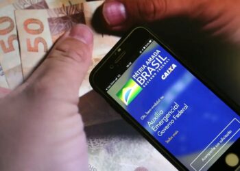 Mãos segurando um smartphone com o aplicativo Auxílio Emergencial da Caixa e cédulas de 50 reais, ilustrando o tema de devolução de valores indevidos.