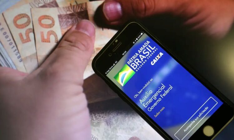 Mãos segurando um smartphone com o aplicativo Auxílio Emergencial da Caixa e cédulas de 50 reais, ilustrando o tema de devolução de valores indevidos.