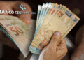 Pessoa segurando notas de reais com o logotipo do Banco Central do Brasil ao fundo.