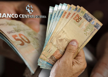 Pessoa segurando notas de reais com o logotipo do Banco Central do Brasil ao fundo.