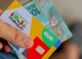 Cartão do Programa Bolsa Família com cédula de 100 reais sendo segurados por uma mão.