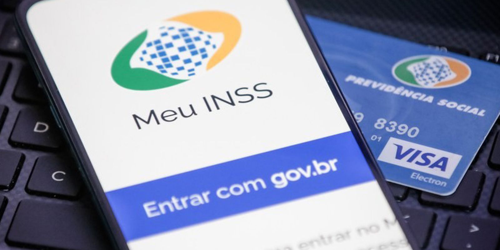 Tela do celular com o aplicativo Meu INSS aberto ao lado de um cartão físico azul da Previdência Social sobre teclado