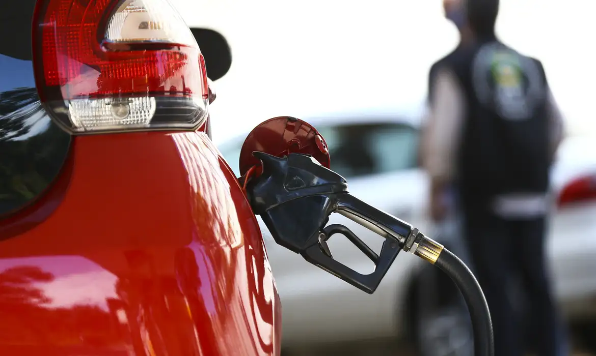 Bomba de combustível abastecendo carro vermelho em posto de gasolina com pessoa ao fundo