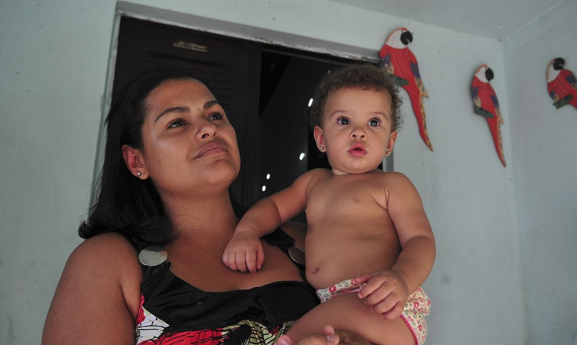 Mulher segura bebê pequeno no colo dentro de casa com parede azul clara e pássaros decorativos vermelhos