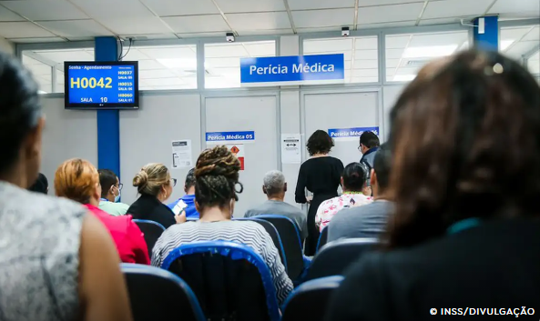 Sala de espera de perícia médica do INSS com pessoas sentadas e um painel indicando número H0042.