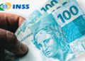 Mão segurando notas de 100 reais com logo do INSS ao fundo.