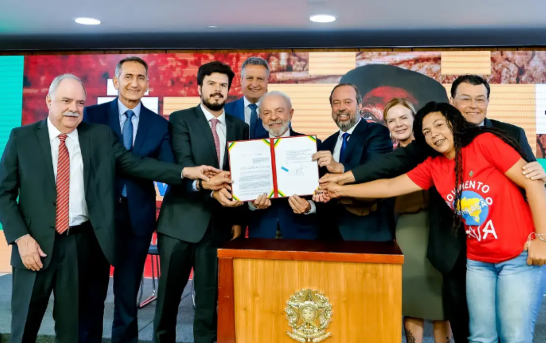 Grupo de líderes e representantes segurando documento oficial em evento formal, com fundo colorido e brasão nacional.