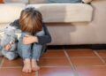 Criança sentada no chão, abraçando os joelhos e um brinquedo de pelúcia, simbolizando a vulnerabilidade de filhos e dependentes de vítimas de feminicídio.