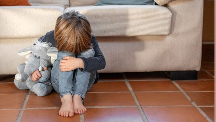 Criança sentada no chão, abraçando os joelhos e um brinquedo de pelúcia, simbolizando a vulnerabilidade de filhos e dependentes de vítimas de feminicídio.