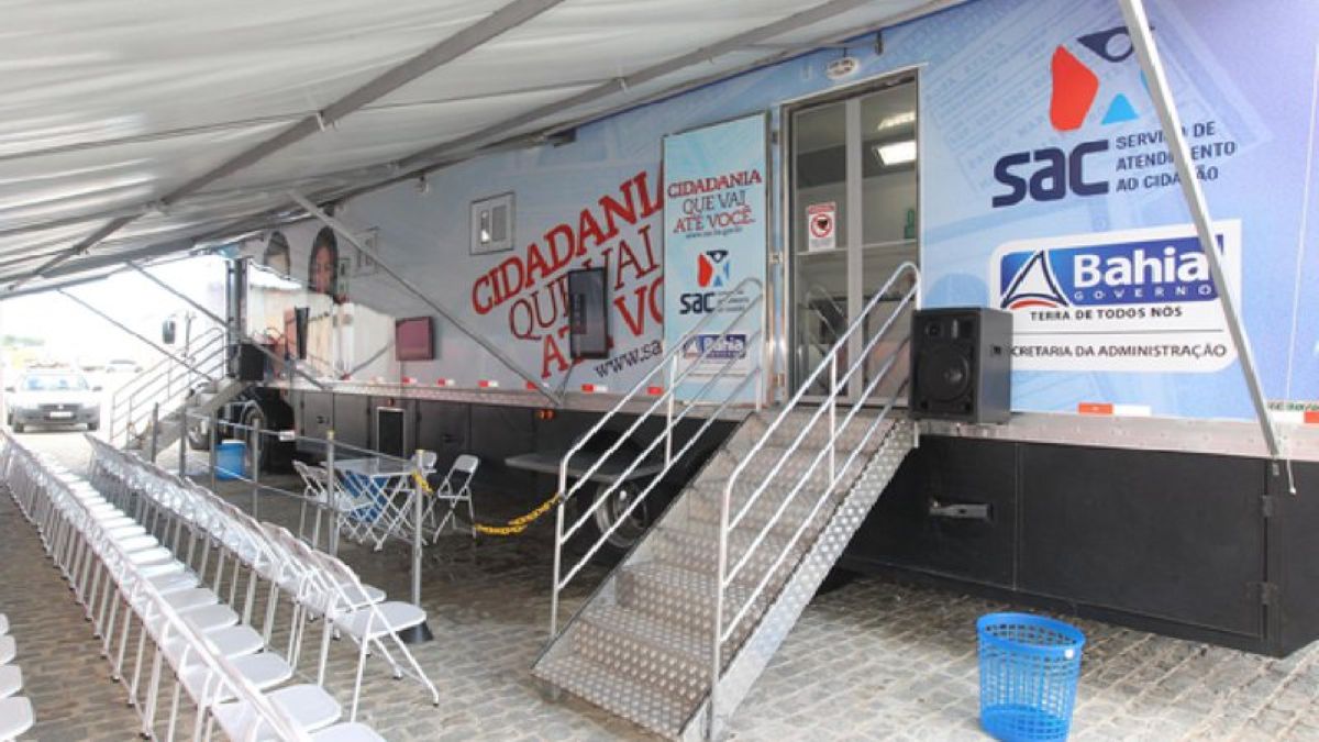 Imagem de uma carreta do SAC Móvel estacionada sob uma tenda, com escadas e cadeiras de espera. A unidade móvel faz parte da estrutura que atende o mutirão da CIN para jovens na Bahia.