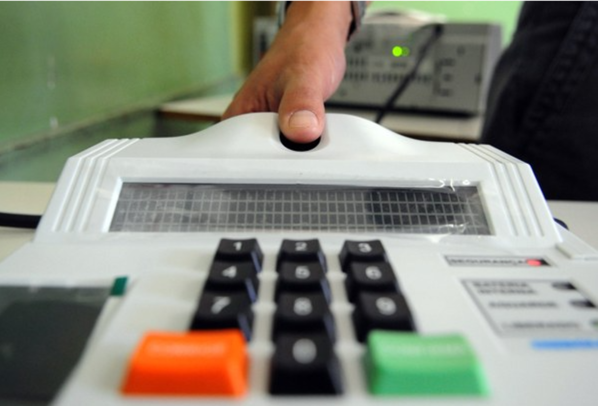 Pessoa colocando o dedo em um leitor biométrico conectado a uma máquina com teclado numérico.