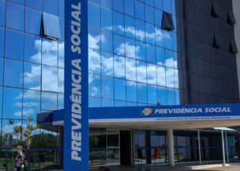 Fachada moderna e espelhada da sede do Instituto Nacional do Seguro Social (INSS), com o pilar azul vertical escrito "PREVIDÊNCIA SOCIAL" em letras grandes, indicando o local para solicitar a devolução de valores descontados indevidamente.