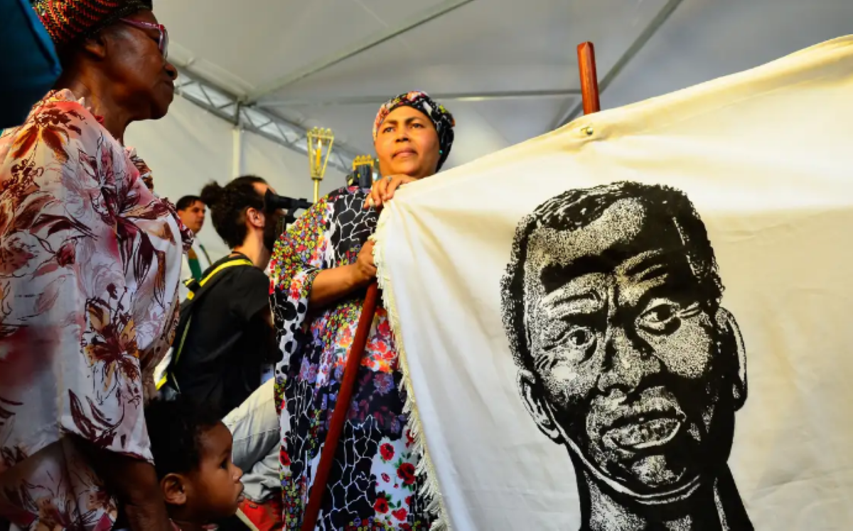 Mulheres segurando um estandarte com ilustração em preto e branco de um homem, durante evento cultural relacionado ao Dia da Consciência Negra.