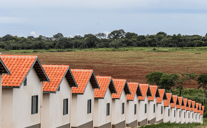 Fileira de casas geminadas brancas com telhado laranja em terreno rural, representando imóveis do programa Minha Casa Minha Vida com limite ampliado para faixas de baixa renda.