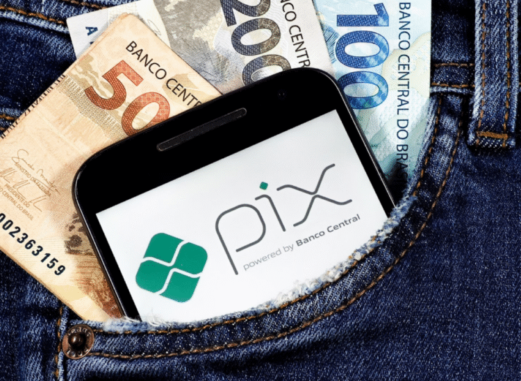 Celular com logo do Pix no bolso de calça jeans junto a cédulas de reais.