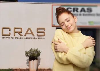 Mulher sorrindo e abraçando a si mesma em frente a unidade do CRAS, representando acolhimento nos atendimentos.
