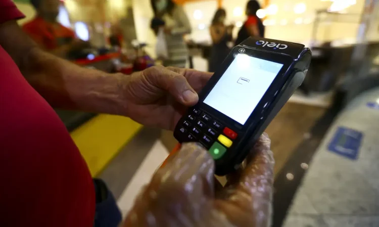 Cartão alimentação de R$ 200 está disponível para inscritos no CadÚnico - Digite seu CPF e descubra se você tem direito 1 Pessoa segurando máquina de cartão com luvas plásticas em ambiente comercial