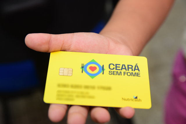 Mão segurando o Cartão Amarelo do Programa Social Ceará Sem Fome, com os logotipos de prato, talher e coração.