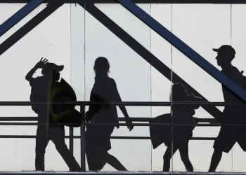 Silhuetas em preto de quatro pessoas caminhando em passarela com estrutura metálica contra fundo claro