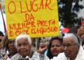 Marcha Nacional das Mulheres Negras do Bolsa Família acontece dia 25 de novembro 2 Pessoas em protesto segurando placa amarela com frase sobre lugar da mulher preta