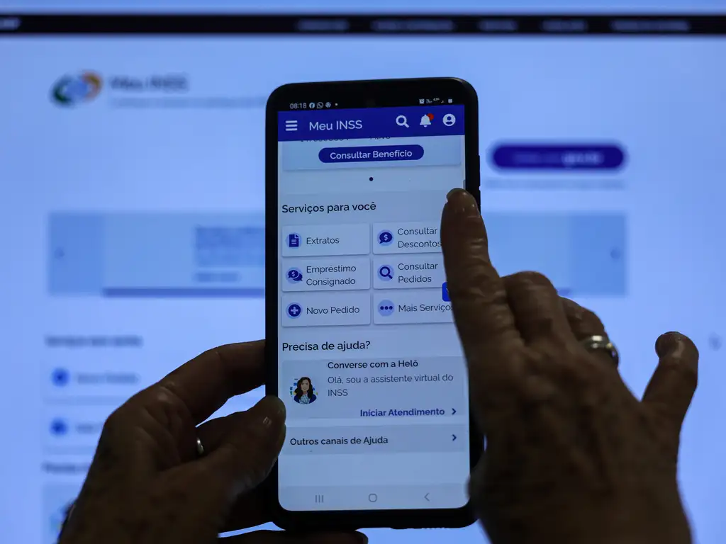 Meu INSS: Portal Digital Seguro para Consultar Benefícios e Serviços da Previdência Mãos utilizando o aplicativo Meu INSS em um smartphone, com o menu de serviços visível, incluindo 'Consultar Benefício' e 'Empréstimo Consignado'.