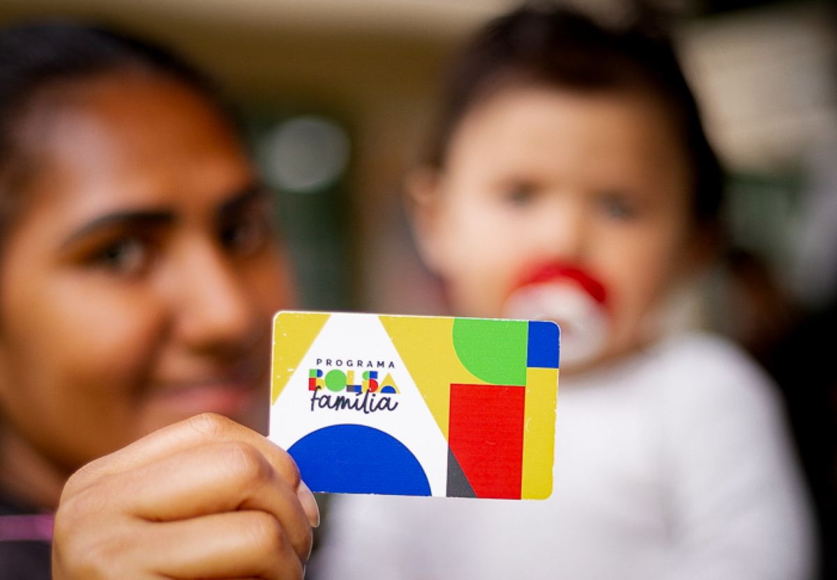 Mulher segurando um cartão do Programa Bolsa Família, com um bebê desfocado ao fundo.
