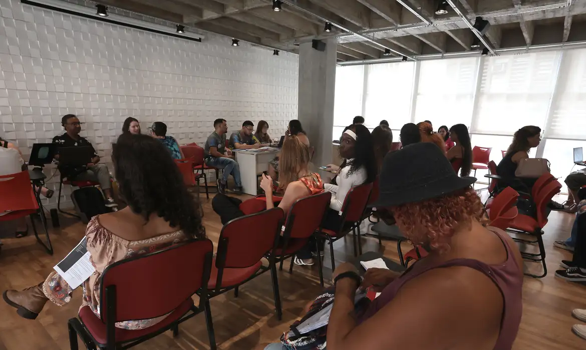 Grupo de pessoas sentadas em cadeiras vermelhas em sala iluminada, interagindo e lendo materiais