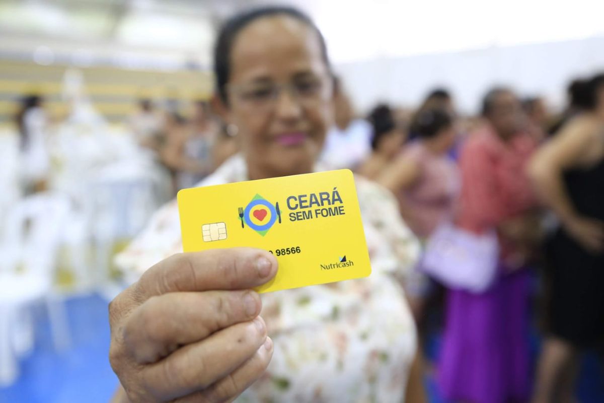 Mulher beneficiária exibindo o cartão "Ceará Sem Fome", com pessoas ao fundo, simbolizando a retirada de documentos.