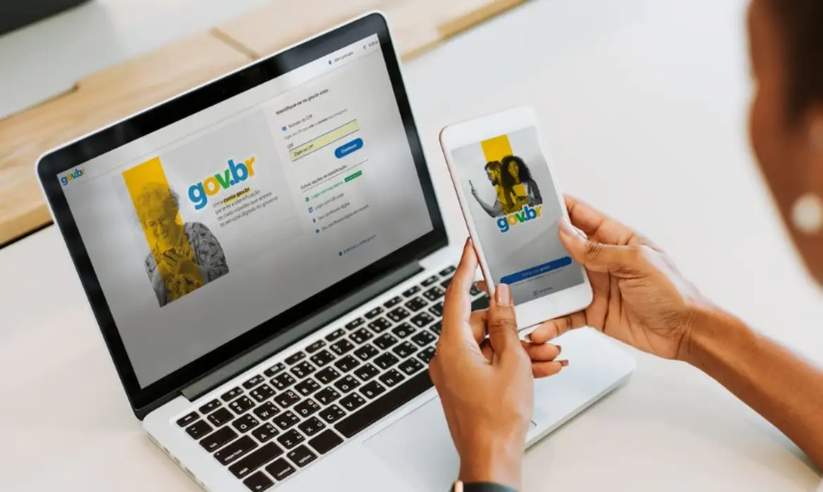 Pessoa usando celular e laptop com telas de login do gov.br em ambiente claro.