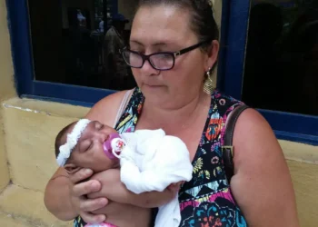 Mulher de óculos segurando bebê dormindo com faixa branca na cabeça e chupeta roxa