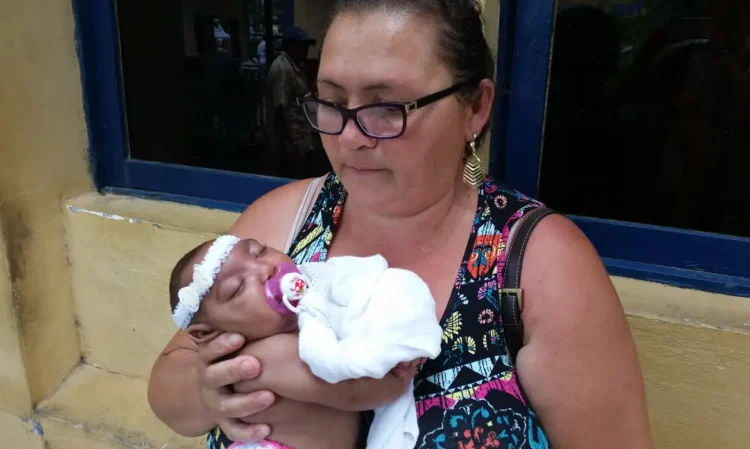 Governo Federal libera pensão vitalícia para famílias de crianças com zika vírus 1 Mulher de óculos segurando bebê dormindo com faixa branca na cabeça e chupeta roxa