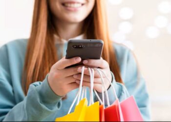 Mulher segurando sacolas coloridas e usando o celular, representando atenção aos golpes durante a Black Friday.