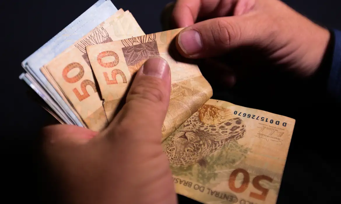 Reajuste Salário Mínimo 2026: Mãos contando dinheiro (cédulas de R$ 50 Reais), representando o novo valor do piso nacional, que deve subir de R$ 1.518,00 para R$ 1.631,00, conforme o PLOA.