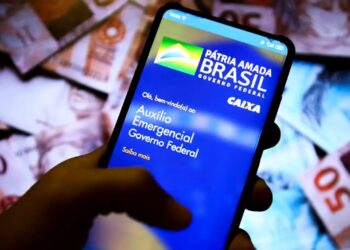 Auxílio Emergencial: quem não devolver até janeiro terá CPF bloqueado e protestado 5 Mão segurando smartphone com o aplicativo "Auxílio Emergencial" na tela, notas de R$ 50 ao fundo, simbolizando a devolução de débitos.