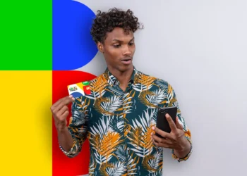Homem com camisa estampada segurando um cartão e olhando o celular com fundo colorido dividido em verde, azul, vermelho e amarelo.