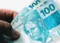 R$103 a mais no seu bolso: veja o novo salário mínimo que começa em janeiro de 2026! 17 Mão feminina segurando cédulas de 100 reais do Brasil com fundo branco