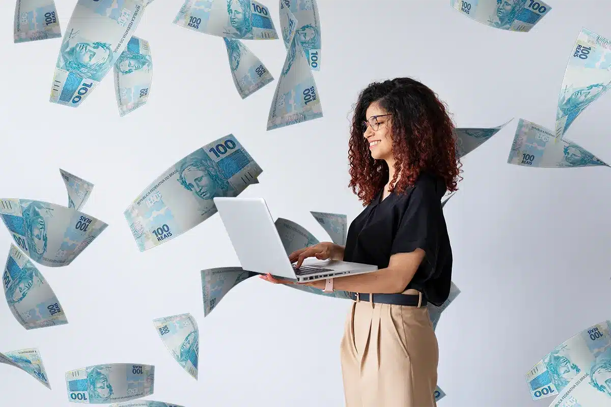 Mulher feliz usando laptop com cédulas de 100 reais voando Mulher sorridente segurando laptop em meio a várias cédulas de 100 reais flutuando ao redor