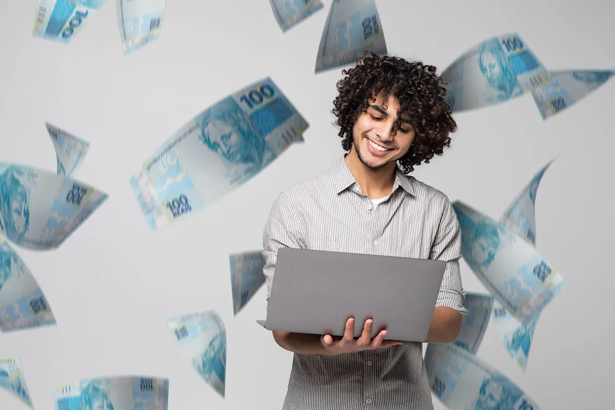 Rapaz sorridente segurando notebook enquanto cédulas de cem reais caem ao redor em fundo cinza