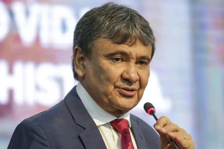 Homem vestido com terno azul e gravata vermelha falando no microfone em evento ao fundo desfocado