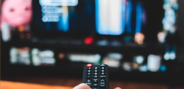 Mão segurando controle remoto com TV ligada desfocada ao fundo