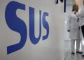 Close na sigla "SUS" em letras azuis sobre uma parede branca de hospital, com profissionais de saúde ao fundo conversando em um corredor iluminado.