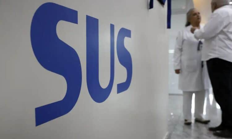 Close na sigla "SUS" em letras azuis sobre uma parede branca de hospital, com profissionais de saúde ao fundo conversando em um corredor iluminado.