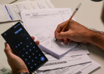 Pessoa usando calculadora no celular e escrevendo cálculos financeiros em bloco de notas sobre faturas