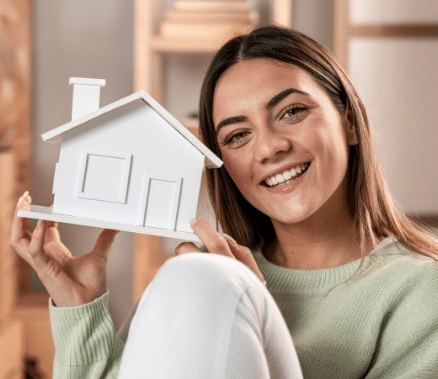 Jovem sorridente segurando uma miniatura de casa branca em ambiente interno